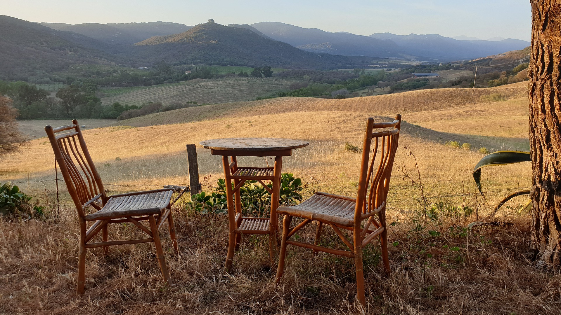 Zwei Holzstühle und ein kleiner Tisch am Rande einer großen Freifläche in der hügeligen Landschaft Korsikas bei angenehmer Abendsonne