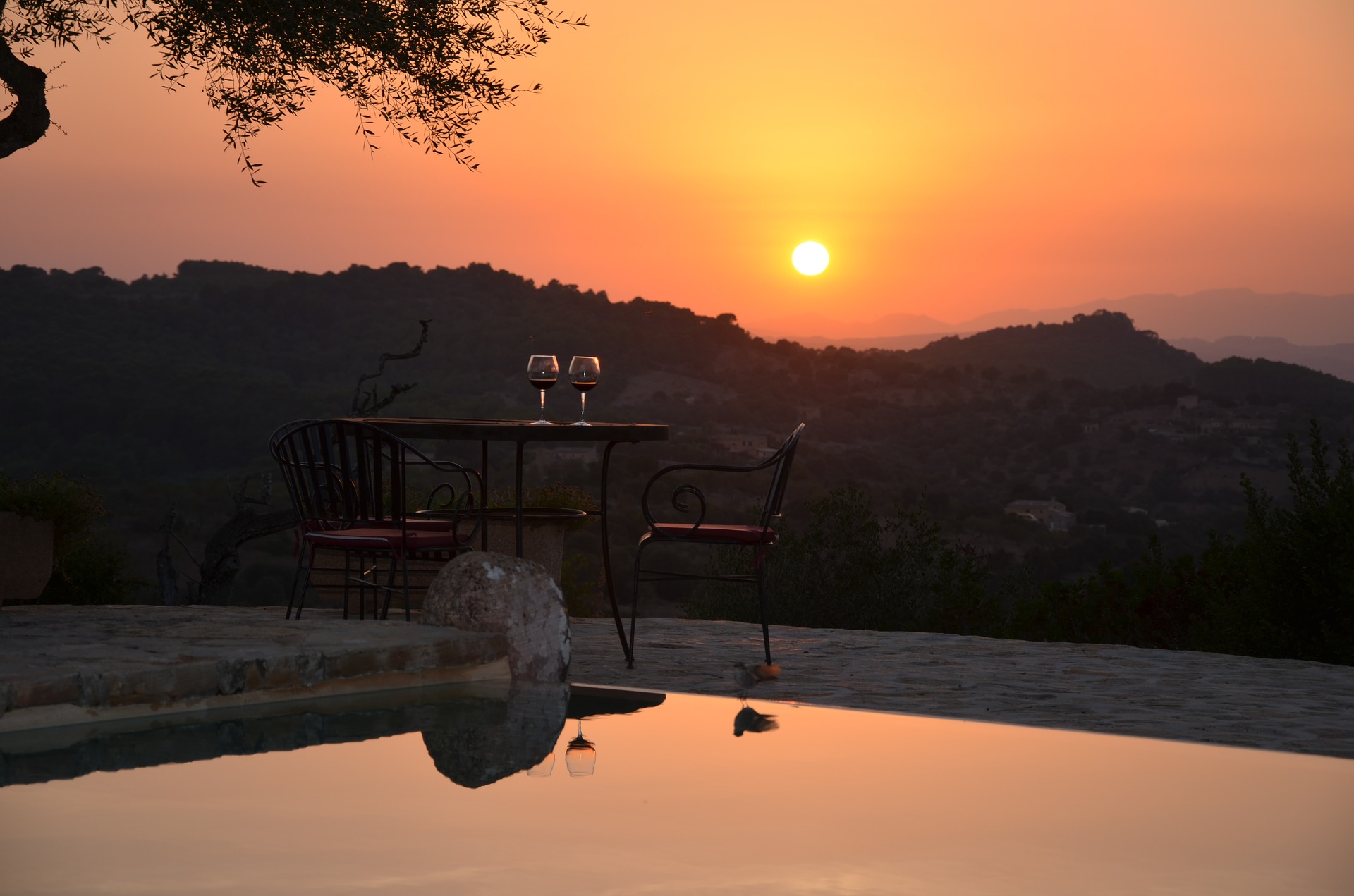 Steinterrasse mit Pool, auf der ein Tisch mit zwei vollen Rotweingläsern steht, im Hintergrund die hügelige Landschaft Mallorcas bei Sonnenuntergang