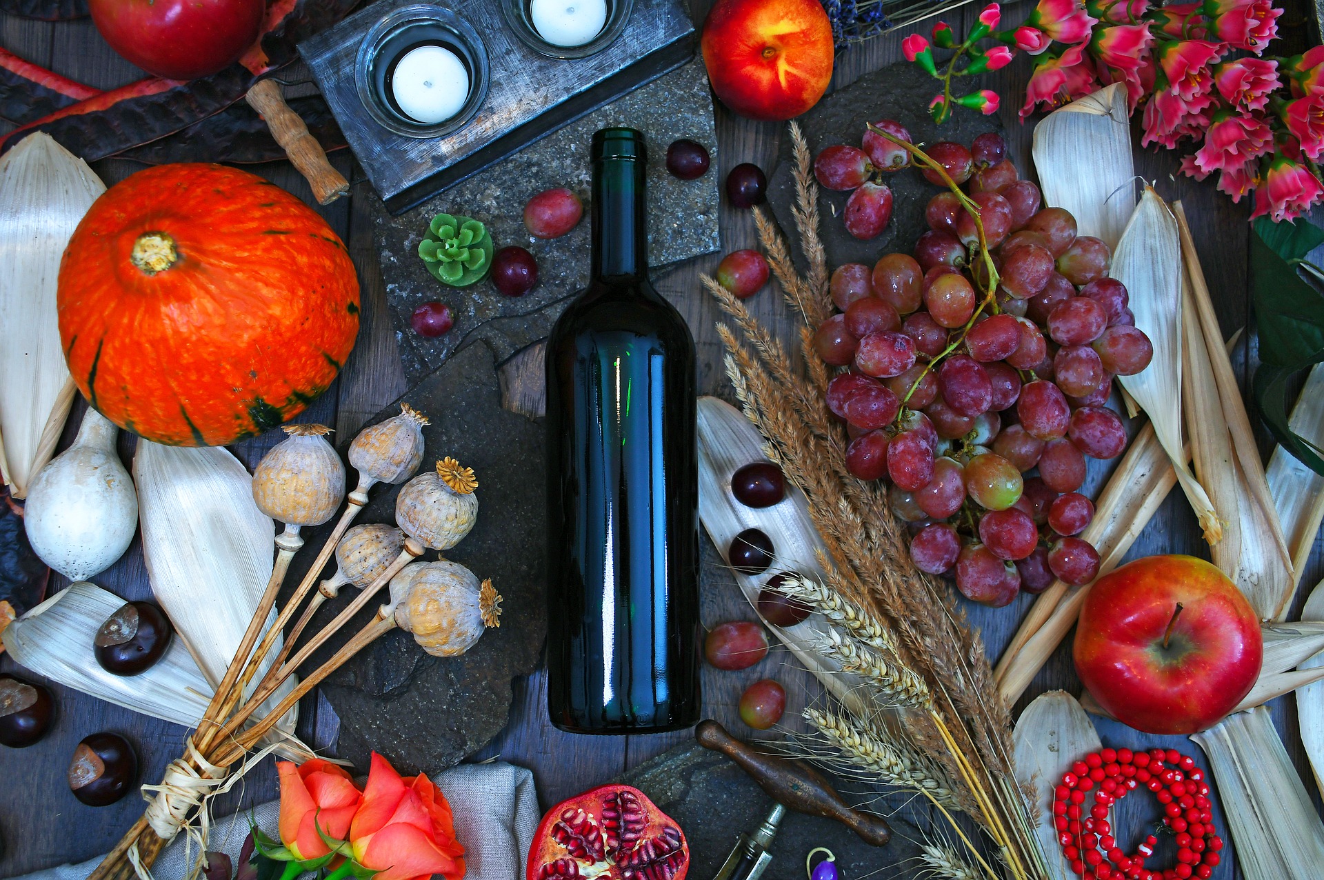 Rustikaler Holztisch, auf dem eine Weinflasche neben Weintrauben, Äpfeln, Knoblauch, Kastanien und Rosen liegt