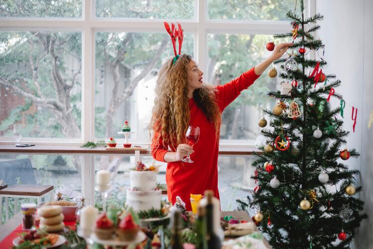 Eine Frau schmückt einen Weihnachtsbaum, hat einen Wein in der Hand und neben ihr stehen Weihnachtsdesserts