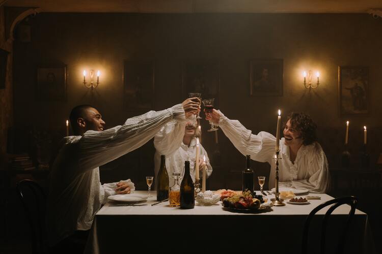 Drei Männer in altertümlicher Kleidung stoßen bei Kerzenschein mit Wein an.