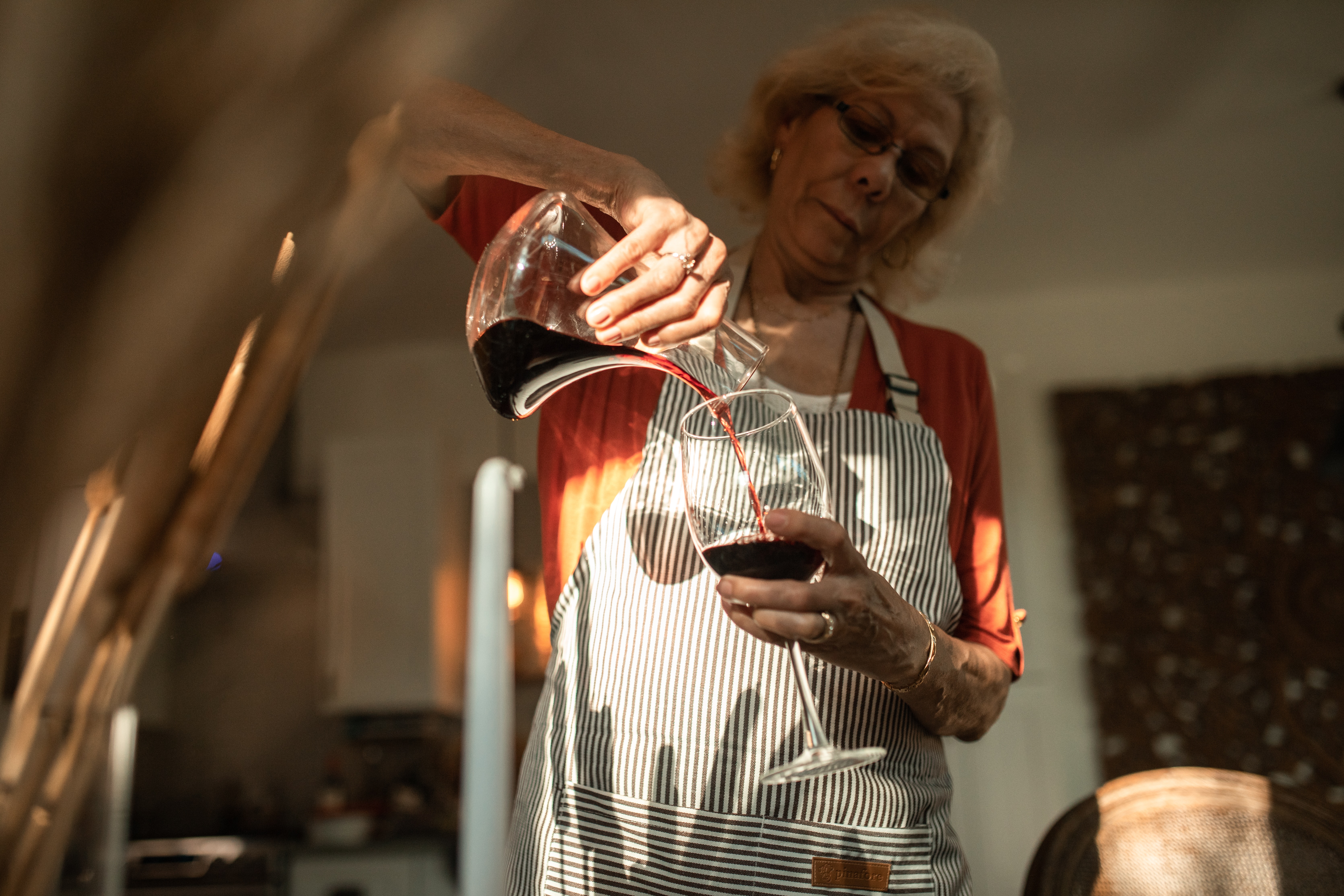 Eine Frau mit gestreifter Schürze gießt Rotwein aus einem Dekanter in ein Weinglas.